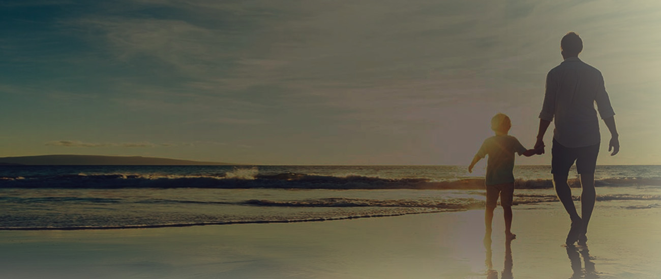 A man and a child walk along the beach, silhouetted against a vibrant sunset sky.