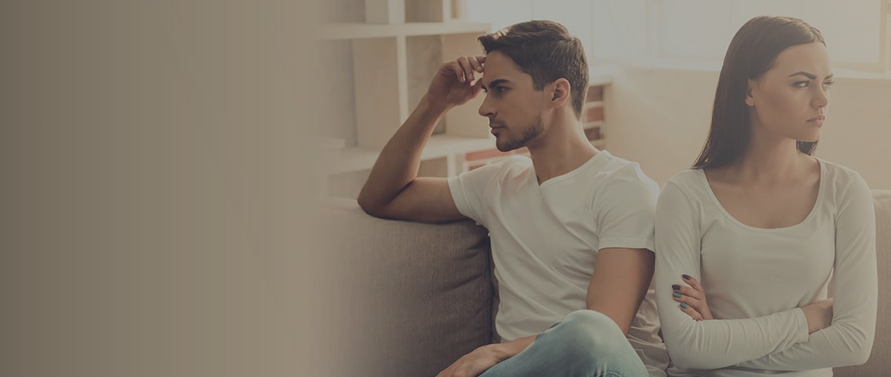 A man and woman sit on a couch, facing each other with angry expressions after a heated discussion.